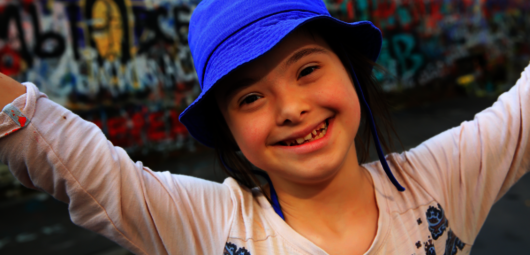 A photo of a young girl wearing a blue bucket hat. She has her arms wide open and is smiling. 